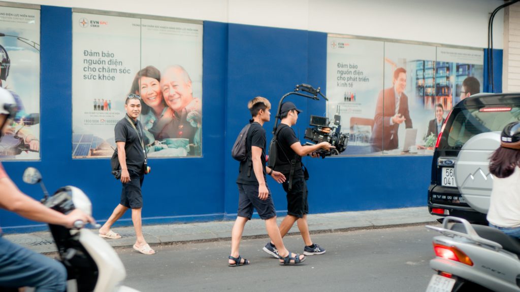 Two men, holding a camera, walking in an alley.