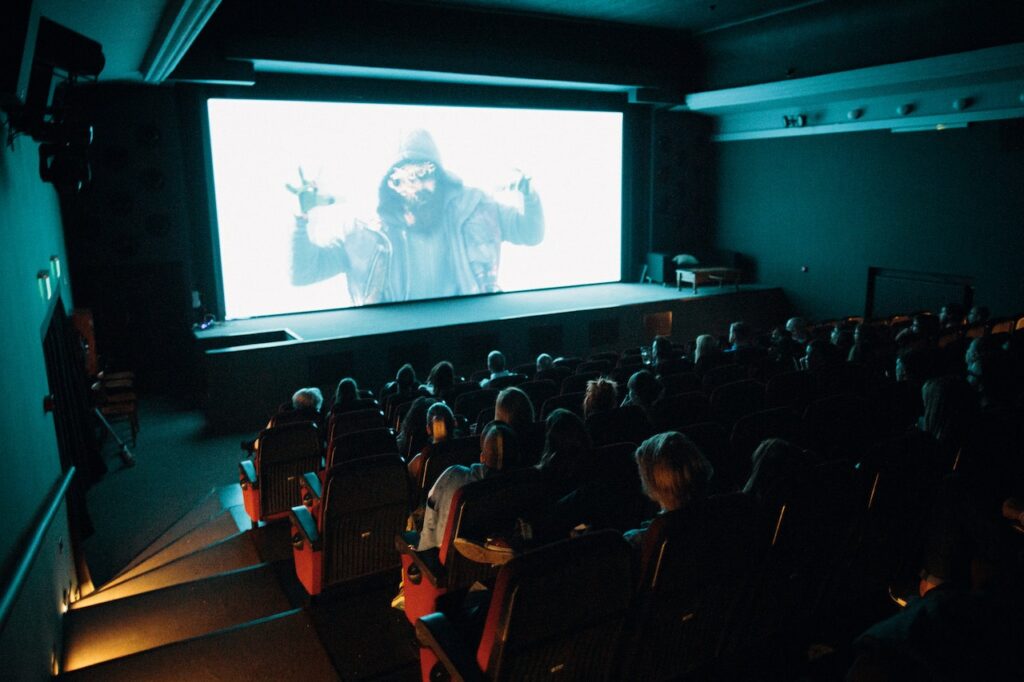 Group of people watching movie in a theatre with man on screen.