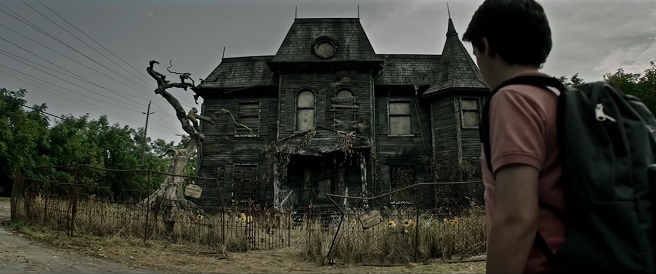 Screenshot from It, where a boy stands outside a run-down house.