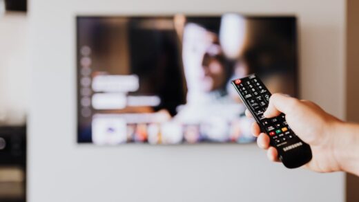 A person holding a television remote in front of their TV.