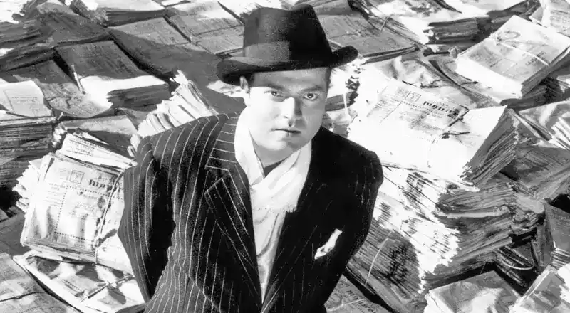 Charles Foster Kane standing atop a piles of newspapers.